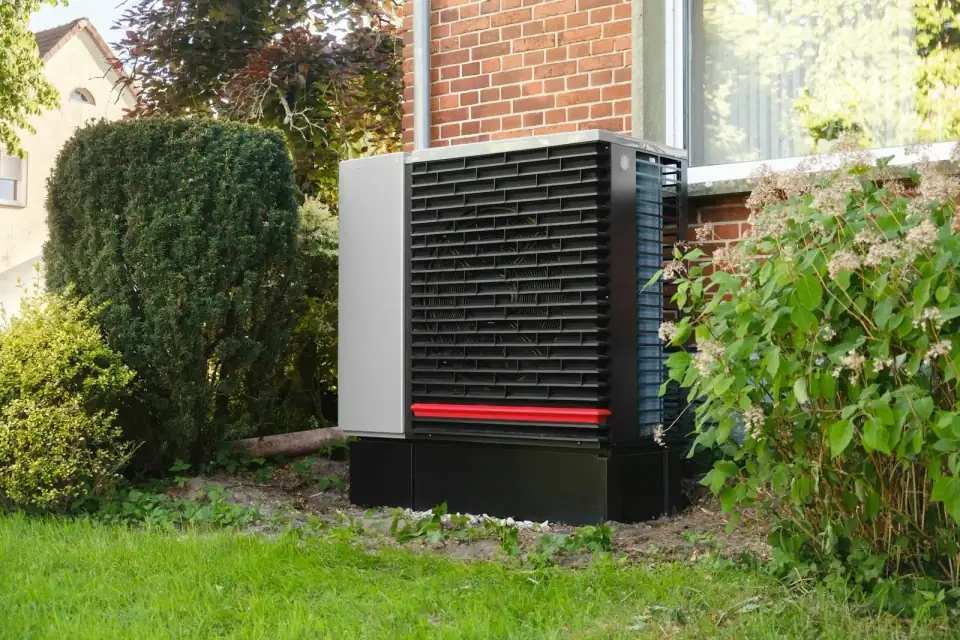 Een buitenunit van een lucht-water-warmtepomp op een sokkel in de tuin, tegen de bakstenen gevel van een woonhuis, tussen een taxushaag en een hortensia