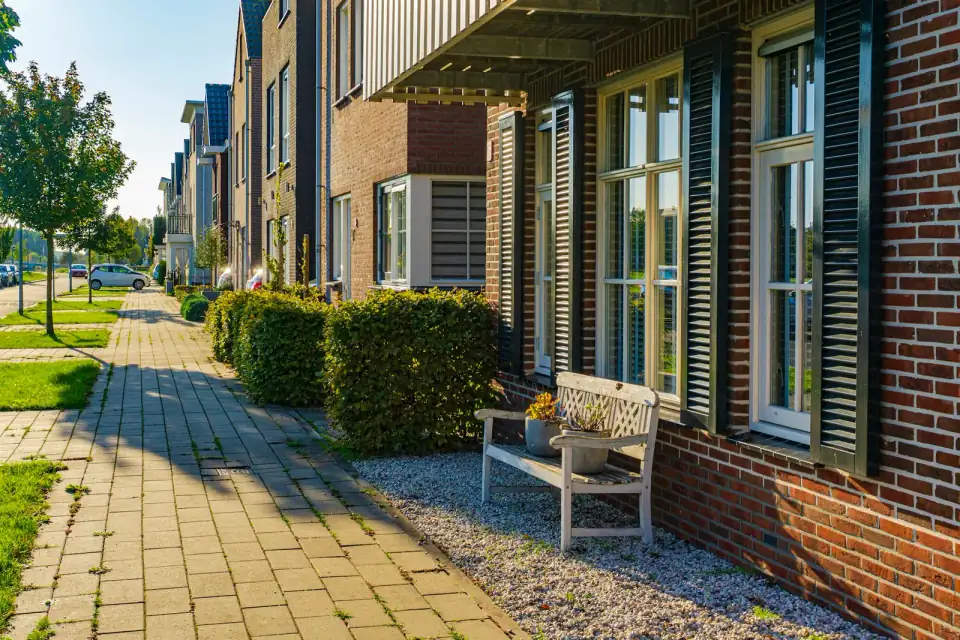 Nieuwere bakstenen rijwoningen met grote ramen, luiken en een bankje aan een stoep met hagen