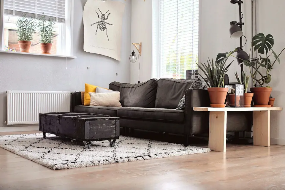 Een woonkamer met een witte paneelradiator onder het raam, een donkere bank, planten in de vensterbank en een bakstenen gevel aan de overkant